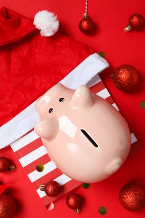 Piggy bank on gift box, balls and Santa hat on red background, top view