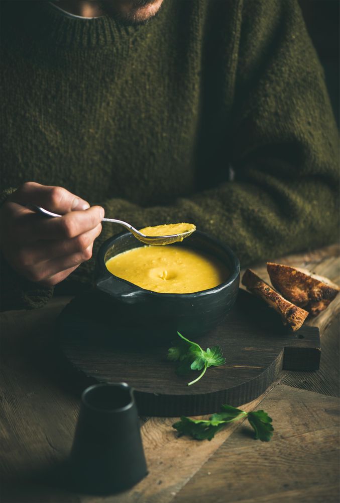 Man with stubble eating warm yellow soup from dark bowl with toast