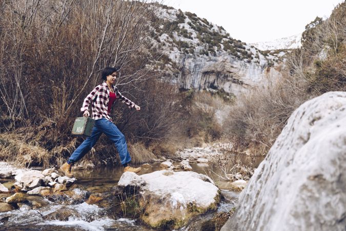 Woman with case jumping on rocks above stream