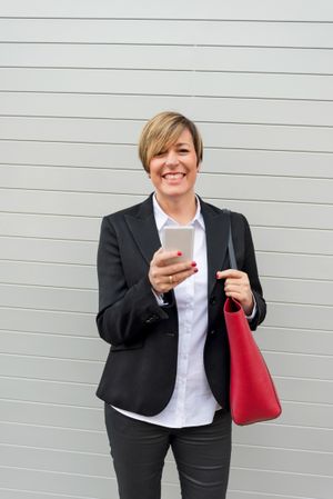 Smiling businesswoman holding out smartphone in front of wall