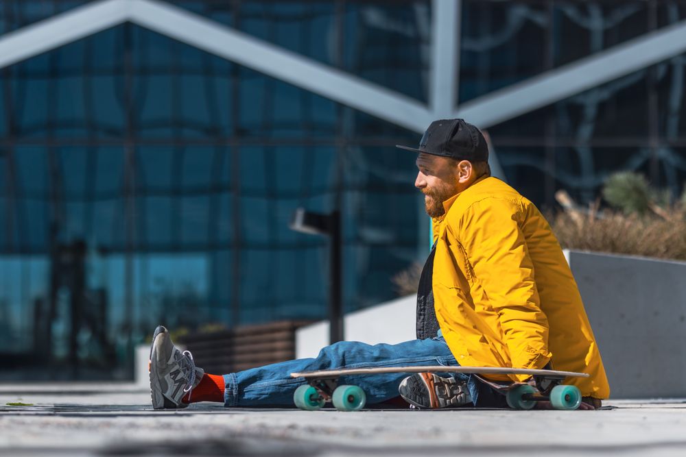 A Young Man In A Vibrant Yellow Jacket And Baseball Cap Enjoys A Sunny Day Skateboarding