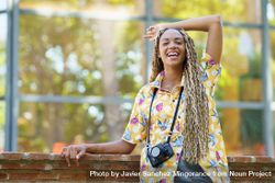 Smiling female in bold patterned shirt leaning on brick wall with camera and arm above head beZgE0