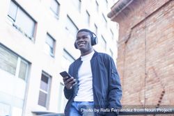 Smiling Black man leaning outside, wearing headphones and holding his phone bYqNJN
