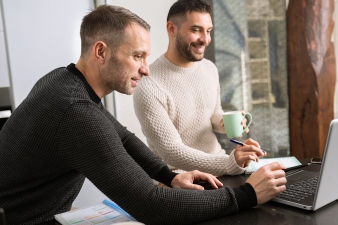 Two men sitting at kitchen counter looking at laptop