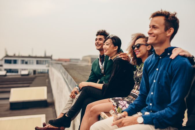 Multi-racial group of  friends sitting on ledge of building on city rooftop