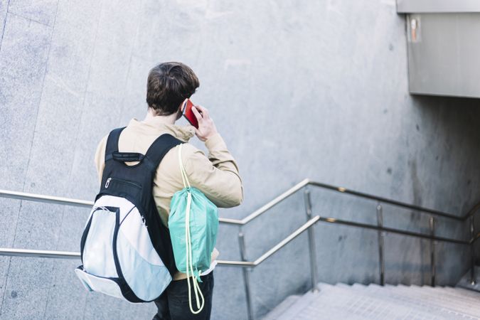 Back of man walking down stairs with backpack while using mobile phone