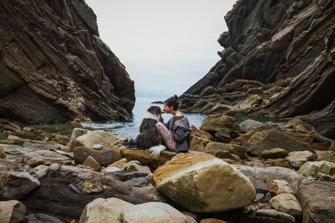 Traveling woman kissing dog sitting on boulder coast