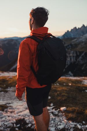 Back view of man in red jacket with backpack standing in the mountains at sunset