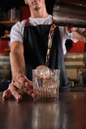 Crop barman pouring drink into glass with ice cubes