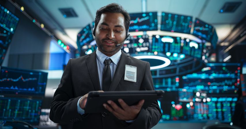 Portrait of a Young Indian Man Working in an International Stock Exchange Company: Happy Trader Using a Tablet Computer, Monitoring, Talking to a Corporate Business Partner in Headphones