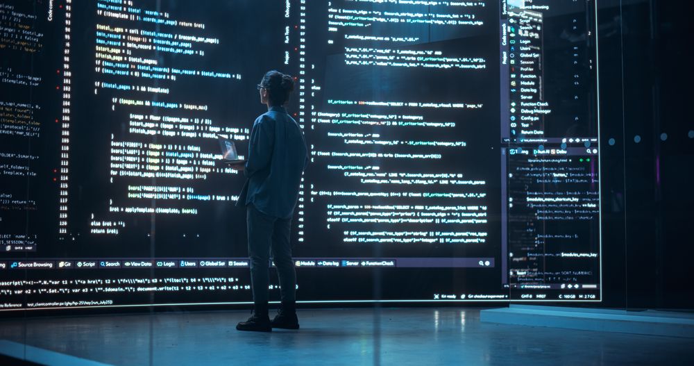 Portrait of Young Woman Working on Laptop Computer, Looking at Big Digital Screen Displaying Back-end Code Lines.