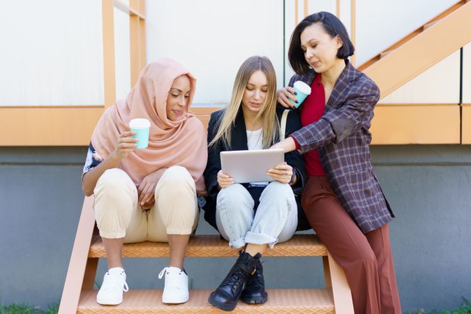 Social female friends sitting on outdoor staircase with coffee and digital tablet
