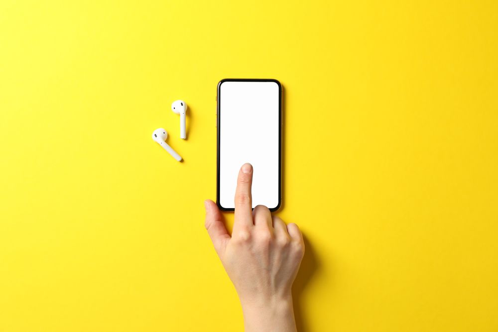 Female hand, phone with empty screen and headphones on yellow background, top view