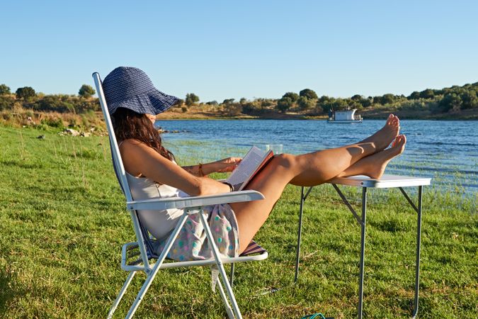 Woman sitting on chair near table and reading book on shore of river