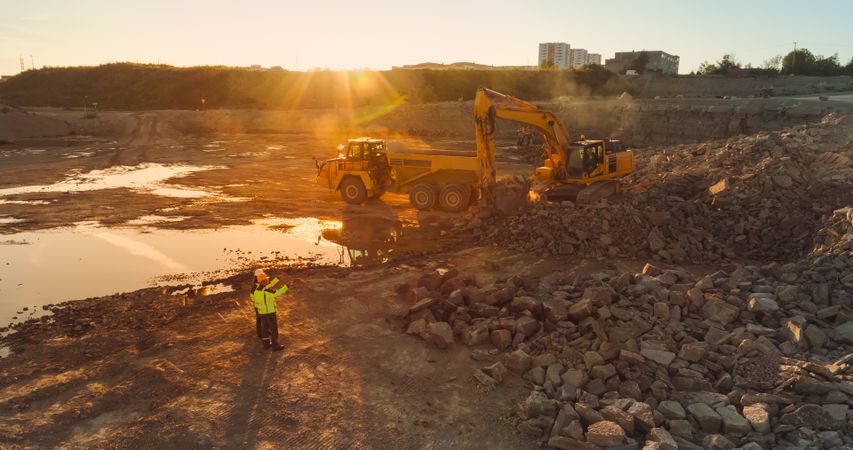 Aerial Drone Shot Of Construction Site On Sunny Evening: Industrial Excavators Digging Rocks And Loading Them Into Truck. Civil Engineer And Inspector Observing Process, Discussing Real Estate Project