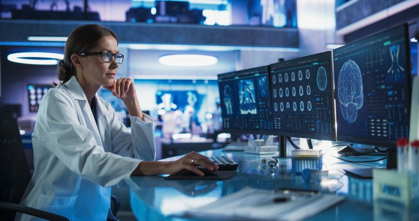 Female Neuroscientist Using Desktop Computer To Analyze MRI Scans Of Human Brain In Busy Research Center Laboratory.