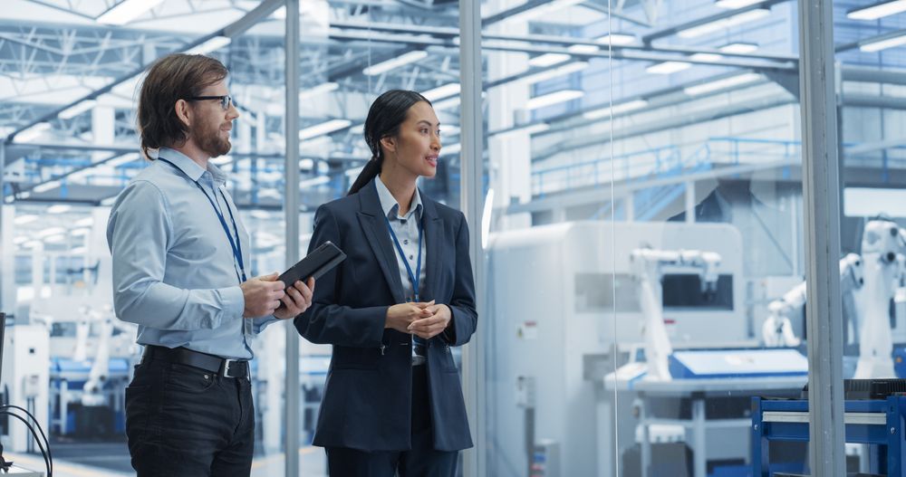 Two Diverse Engineers Walking in Office, Using Tablet Computer at an Electronics Factory. Female and Male Specialists Chatting, Monitoring Robots on Modern Assembly Line.