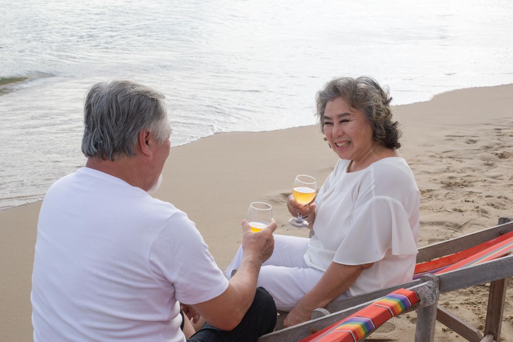 Happy mature Asian couple relaxing on the beach with wine