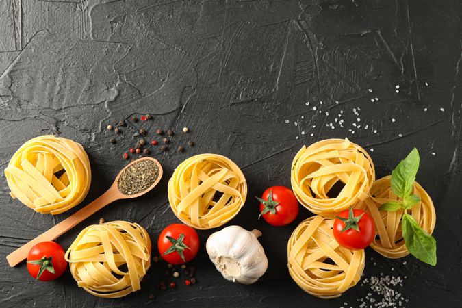Flat lay composition with pasta, tomatoes, salt, garlic and spices on dark background, space for text