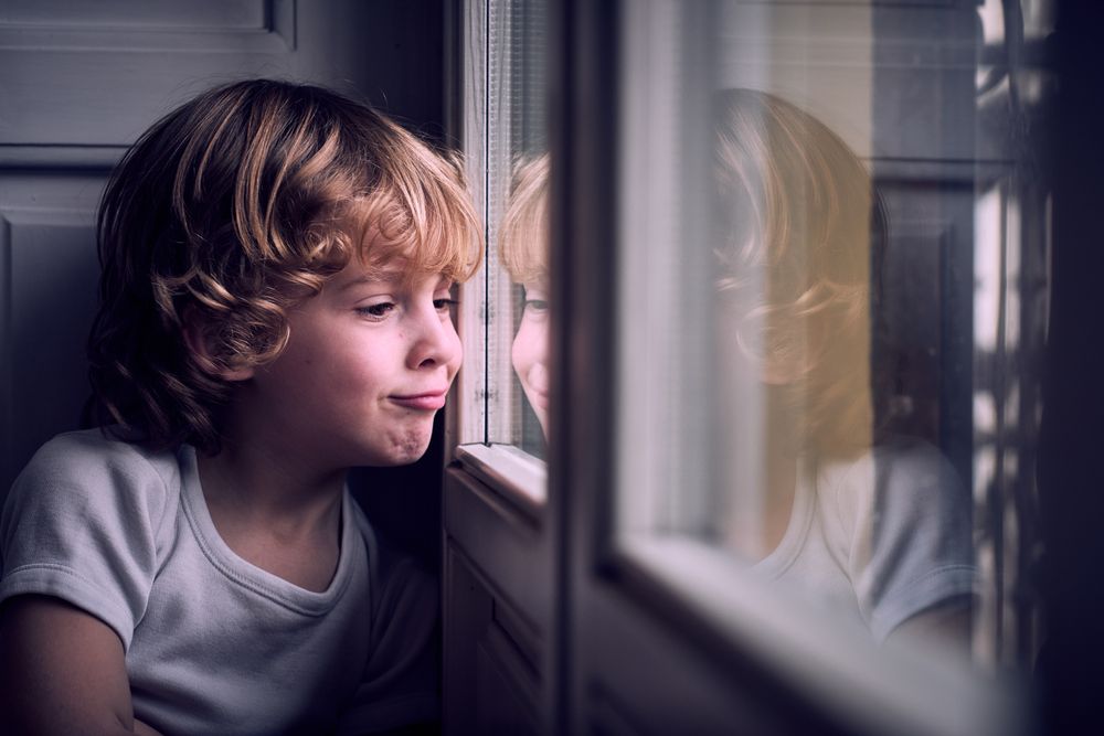 Cute child making face and looking through window at home