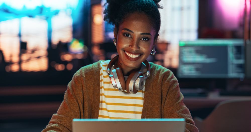 Close Up Portrait of a Young Black Woman with Headphones, Looking at Camera, and Smiling in a Creative Office with Neon Lights.