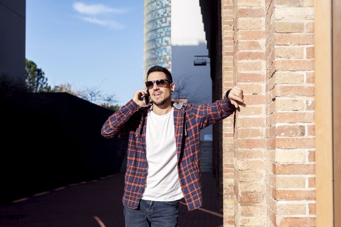 Man standing by brick wall wearing sunglasses and chatting on smartphone outdoors