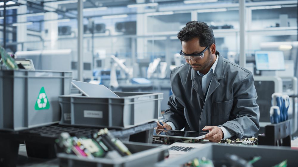 Indian Male Worker Disassembling Laptops To Recycle Electronic Components For Microchip Production At an Automated Electronics Factory With Robotic Arms.