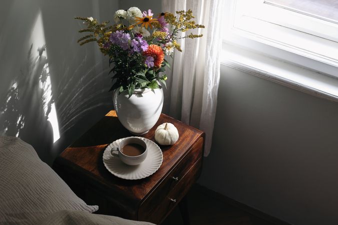 Cup of coffee, pumpkin on wooden bedside table with vase in sunlight