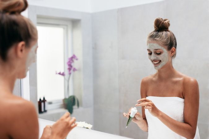 Happy young woman applying face mask in bathroom and smiling