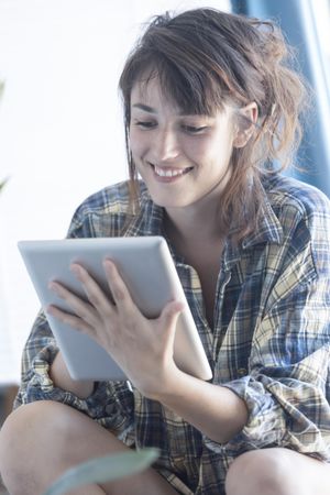 Smiling female in flannel shirt lounging in bright room with digital tablet