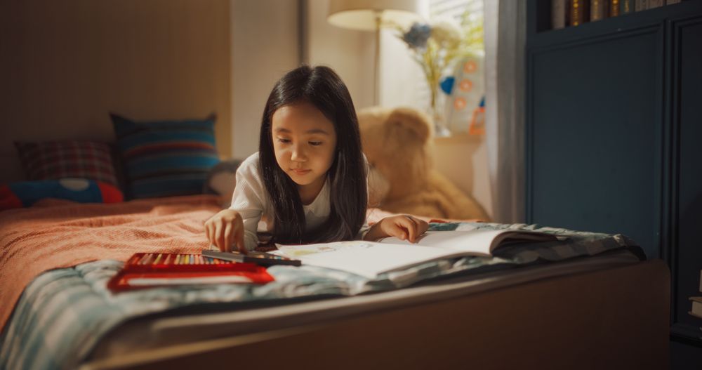 Portrait of a Happy Little South Korean Girl Focused on Designing Her Comic Book with Colorful Pencils While Laying on a Bed in Children's Bedroom.