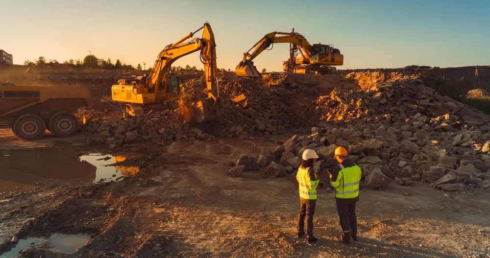 Aerial Drone Shot Of Construction Site On Sunny Evening: Industrial Excavators Digging Rocks And Loading Into A Truck. Civil Engineer And Inspector Observing Process, Discussing Real Estate Project
