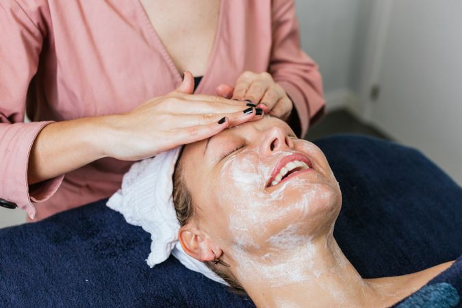 Crop beautician cleansing face of woman in salon