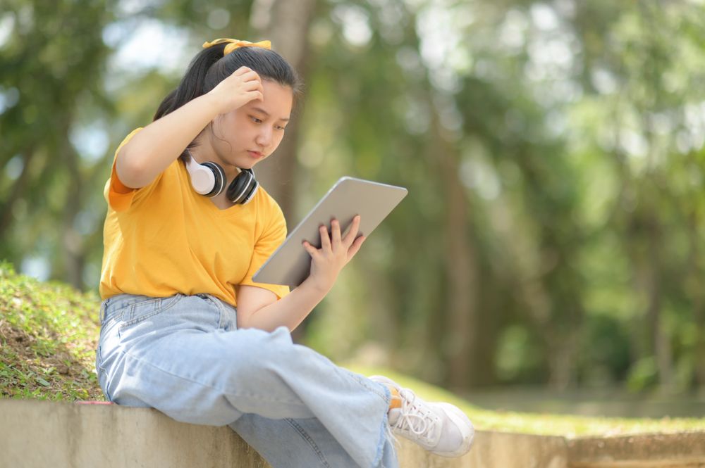 Woman in yellow shirt wearing headphones on neck holding tablet sitting in garden serious pose.