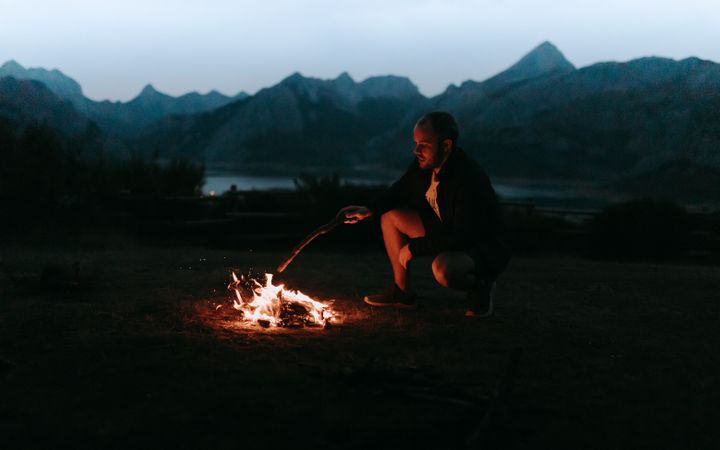 Man sitting beside bonfire in the woods near mountains and lake
