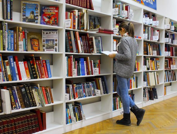 Young woman in jeans standing beside bookshelves at the library