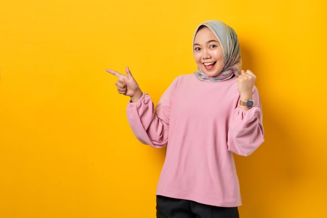 Happy Muslim woman in headscarf pointing to space beside her and raising fist in celebration