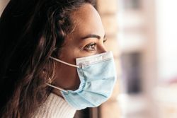Young woman wearing face mask looking out the window at home
