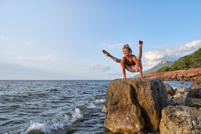 Cheerful sportswoman taking Tittibhasana pose on rock near sea
