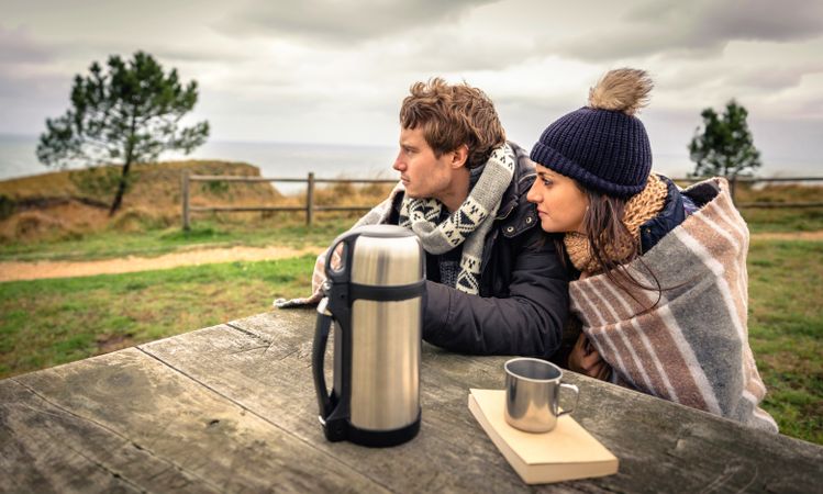 Couple wrapped in blanket on cold park bench with coffee flask looking in the distance