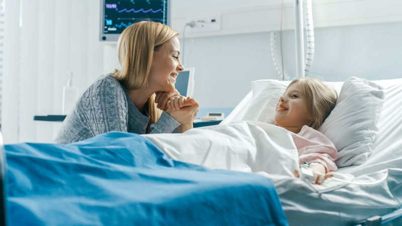 Cute Little Girl Lies on a Bed in the Children's Hospital, Her Mother Sits Beside