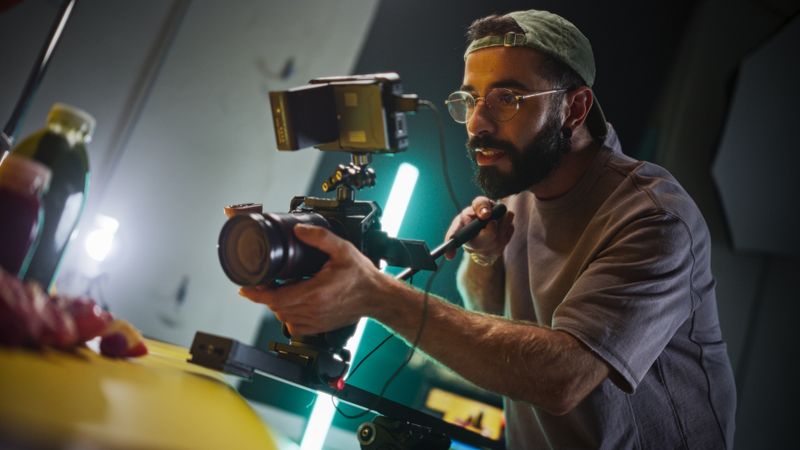 Focused Male Cinematographer With Beard And Glasses Operates Professional Camera On Film Set, Capturing Dynamic Shots Under Vibrant Studio Lights, Showcasing Videography Techniques And Equipment.
