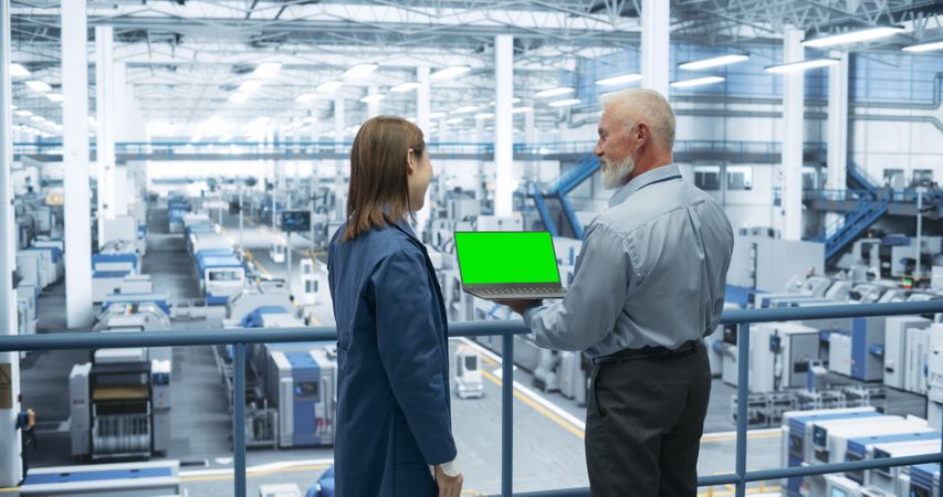 Two Multiethnic Engineers Standing on a Platform, Using Laptop Computer with Green Screen at an Electronics Factory. Machines are Undergoing Maintenance, Specialists Monitoring the Progress
