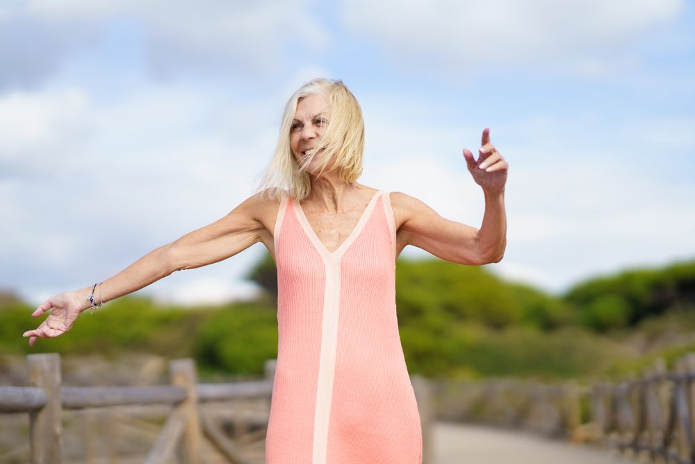Joyful older woman with grey hair dancing on wooden walkway near the coast, copy space