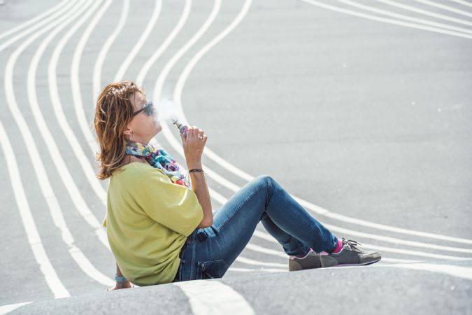 Modern relaxed woman vaping while sitting on marked road