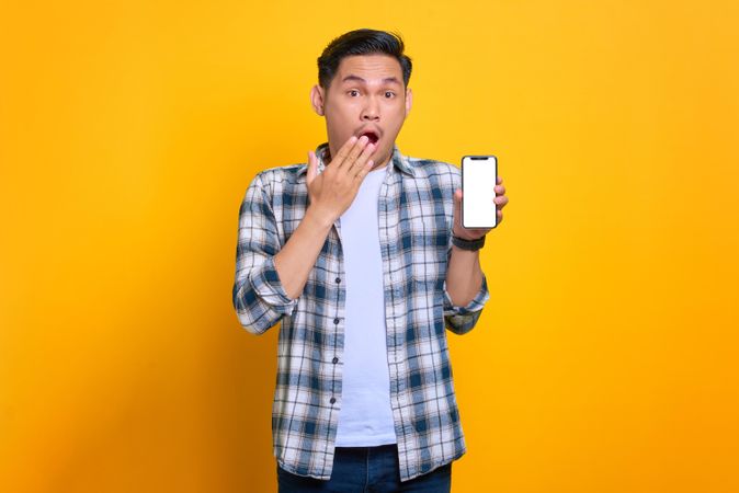 Surprised Asian male with hand over mouth and showing blank screen of smart phone