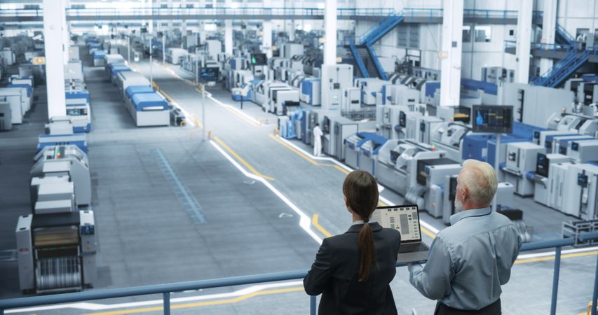 Male Engineer and Female Project Manager Standing on a Platform, Talking, Using Laptop Computer and Monitoring Production at a Modern Automated Electronics Manufacture with AI Robotic Arms