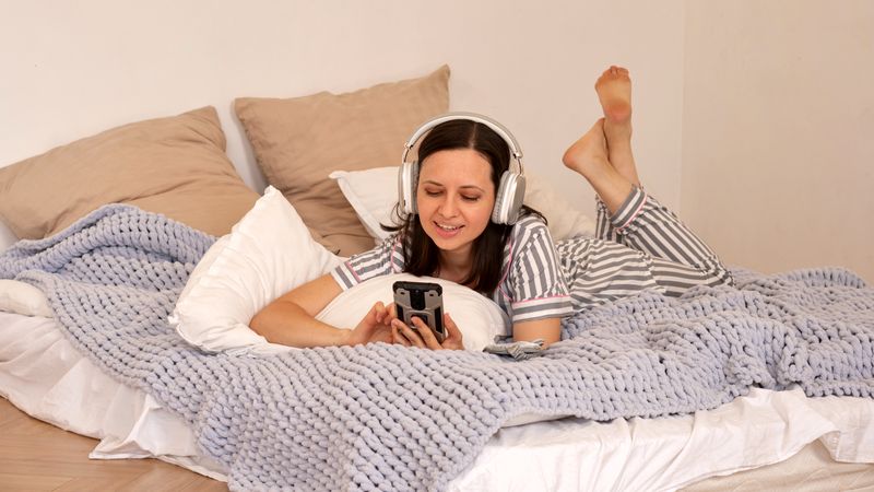 Woman lying in bed with headphones, using smartphone.