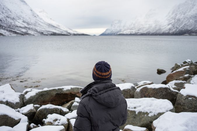 Solitary tourist on rocky coast against calm lake water and snowy mountains in cold overcast weather