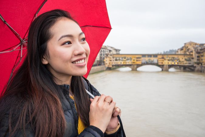 Cheerful Asian tourist with umbrella standing on bridge near river